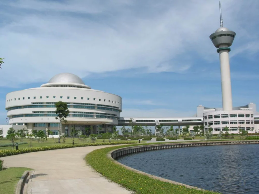 Modern university campus with a domed academic building, tall observation tower, and landscaped lake.