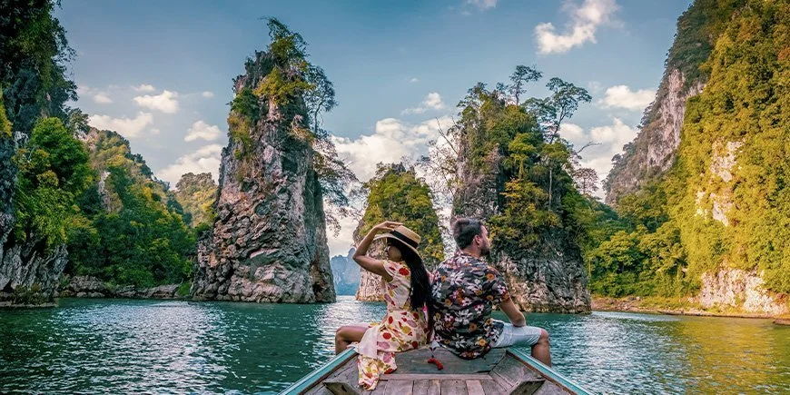 Couple sitting on a wooden boat surrounded by tall limestone cliffs and lush green nature in Thailand.