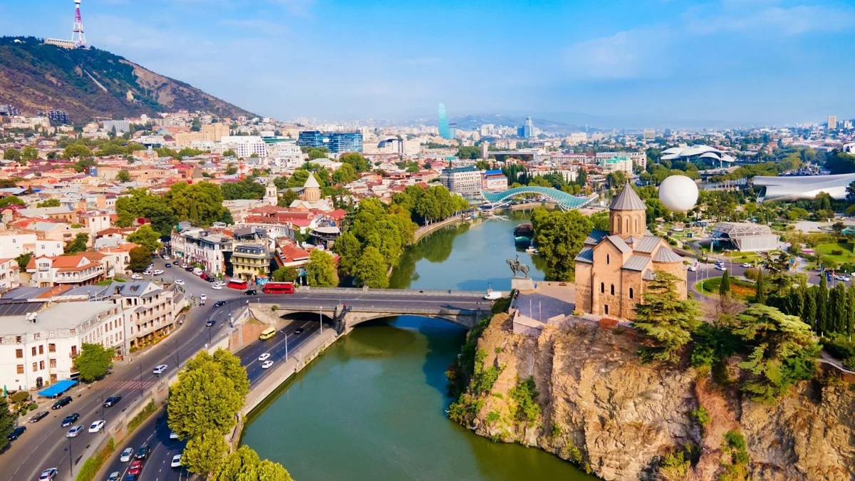 Aerial view of Tbilisi, Georgia showing the Kura River, Metekhi Church on a cliff, historic old town buildings, and the modern Peace Bridge.