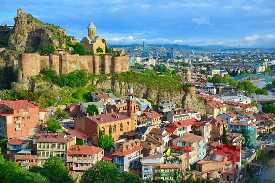 Panoramic view of Tbilisi, Georgia featuring Narikala Fortress, colorful old town houses, the Kura River, and the modern Peace Bridge.