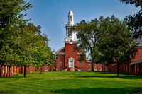 A scenic view of a historic Yale University campus building with red brick architecture, a white clock tower rising above the trees, and a wide green lawn lined with shaded walkways under a clear blue sky.
