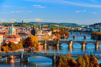Panoramic view of Prague with historic bridges crossing the Vltava River, colorful autumn trees, and classical red-roofed buildings stretching across the cityscape.
