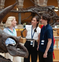 Three students standing beside a dinosaur skeleton exhibit inside a natural history museum.