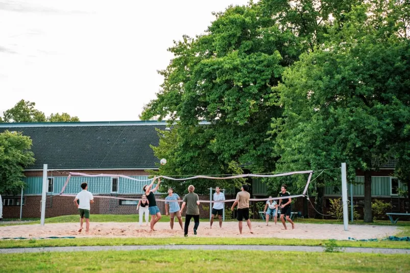 Students playing beach volleyball outdoors on a sunny day surrounded by trees and campus buildings.