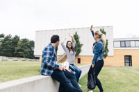 Three university students socializing outdoors on a modern campus courtyard, with two seated on a concrete bench and one standing while waving playfully, surrounded by green lawns and academic buildings.