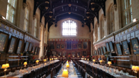 A grand historic university dining hall with long candlelit tables, high vaulted ceilings, ornate wooden details, and large stained-glass windows creating a warm academic atmosphere reminiscent of Oxford college traditions.